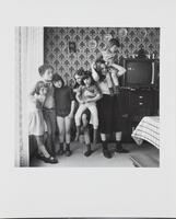 Black and white image of seven children with a television in the background. Two smaller children are held by two taller children and three others have their arms around each other.