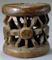 This is a wooden stool with a circular base and a flat, circular seat. The seat is supported by an openwork design consisting of a circle with eight lines projecting outward. This motif is repeated six times around the stool. 