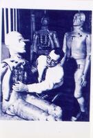 A portrait image in two tones of blue printed slightly off match, showing a man at the center dressed in a white shirt, tie and slacks, with dark-rimmed glasses. The man is leaning over a automoton/robot, which is seated to the left of him. On the right and to the back, are two more standing robots. Each robot has humanoid features like a face, torso, arms, hands, legs, and feet with boots on. Two of the robots, one seated and one to the back at the center, have their internal workings revealed with their central torso panel removed, showing wires and indeterminate parts. 