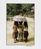 An image of three children walking on a beach under an umbrella, their backs to the viewer. A fourth child stands, out of focus, in the background among buildings.