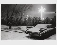 A sedan parked in an empty lot on a snowy night is illuminated by a brightly-lit electric Christmas star.