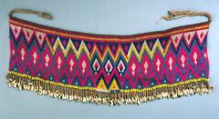 An apron made from multi-colored beads in a geometric pattern with zig-zag and diamond shapes. There are two strings attached at the top of the apron, to tie around the waist. At the bottom of the apron is a fringe of cowrie shells. 