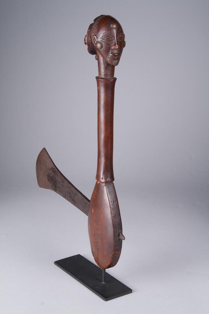 This Hemba <em>adze</em>, or chief’s ceremonial ax, is decorated with an elegantly carved female head upon the end of its smooth handle while an iron blade has been lodged into its oval base. This <em>adze</em> exhibits the characteristic hallmarks of a Southern Hemba style, which in turn was strongly influenced by the neighboring Luba. The head bears an elongated, oviod-shaped face, a wide convex forehead, coffeebean-shaped eyes within ocular recesses, a triangular nose, and full lips. An elaborate pulled-back hairstyle in the form of a chignon (<em>kibanda</em>), features a cruciform motif. Four brass tacks that have been inserted into the extreme top, bottom, left and right points of the face echo this crucifix shape.