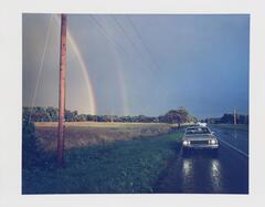Image of cars parked on the side of a country road opposite a double rainbow. 