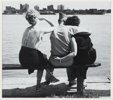 An image of a man and two women sitting on a park bench, seen from behind. The man sits in the middle with his arm around one woman and the other woman has her elbow on his shoulder.