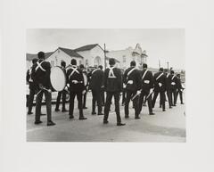 Black and white image of the backs of boys in uniforms. Some of the boys are carrying drums and drumsticks. 