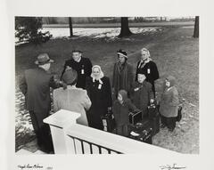 A black and white image of three children and four adults with suitcases greeting two other adults with a white fence in the foreground.