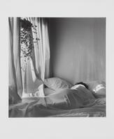 Black and white image of a person lying in a bed under a sheet beside a window in which a plant hangs.