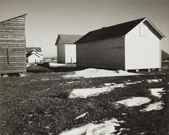 A black and white photograph of several wood-sided buildings. The two on the left are painted white. A white house is visible in the background. The foreground is snow and dirt.
