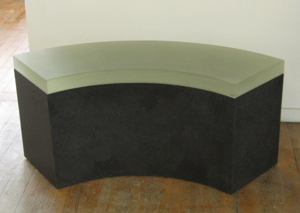 Curved bench with dark brown granite base and curved, frosted or fritted, pale green glass on top.