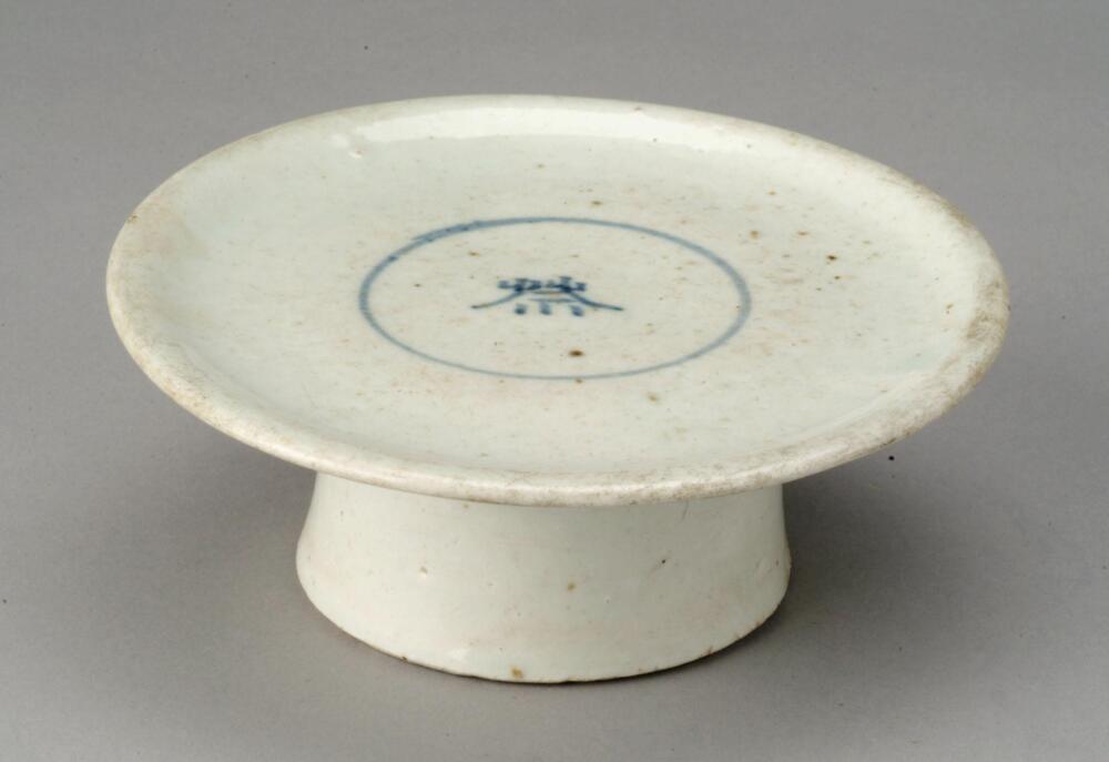 The upper surface of this vessel features a circle with the Chinese character &quot;je (祭: ancestral rite)&quot; rendered inside in cobalt blue pigment. The tray features blemishes, while the rims show traces of use. The foot retains traces of coarse sand supports stuck to it during firing. This type of ritual vessel has been excavated from the upper sediment layers of waste deposits of kilns in front of what is now Bunwon-ri Elementary School in Gwangju-si, Gyeonggi-do. Such vessels are presumed to have been produced immediately before the Bunwon-ri kiln cloised down and to have been widely supplied to the general public.&nbsp;<br />
[Korean Collection, University of Michigan Museum of Art (2014) p.196]
