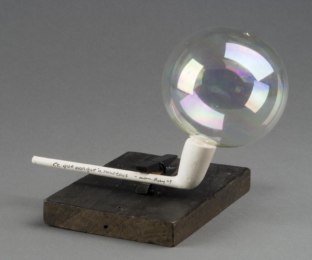 A clear glass bubble rests on the bowl of a plain white clay pipe. Along the stem of the pipe are the words, "Ce que [sic] manque à nous tous."