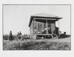 A black and white photograph of a house, single level, positioned to the right of the shot. Three children stand to the left, holding tires, and four more are on the porch.