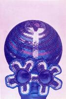 This color photolithograph is in shades of purple and blue and has an image of a woman's head. She is wearing an elaborate hat and glasses. The hat is a large sphere and is covered in stripes of colored sequins. The glasses also have colored sequins but in the shape of flowers.