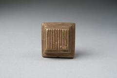 Gold-weight in the shape of a square base with two smaller squares stacked on top, decorated with incised lines. 