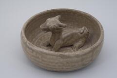 A shallow basin with flat bottom and gently curved sides and a single reclining goat sculpted in the center of the base.&nbsp;