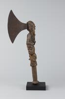 Axe with wooden handle. The bottom of the handle is cylindrical with a small disc-shaped grip at the end, while the upper portion of the handle is composed of two figures on top of one another. They face opposing directions; the lower figure appears to be holding a knife while the other figure may be holding a musical instrument to its mouth. 
