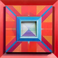 A brightly painted wood frame with an angled surface containing a canvas painted with bright geometric designs of blue, pink, orange, and red. In the center of the canvas is a wood element with the appearance of stairs painted purple, green, blue and yellow.