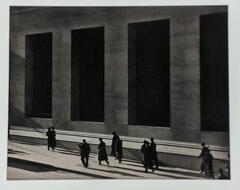 Black and white image of small figures wearing coats, hats, and jackets with their features obscured walking beside a large building. The figures are dwarfed by angular architectural features, either windows or columns, through which only dark spaces are visible.&nbsp;