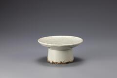 This is a ritual dish assumed to have been produced at the private kiln near Bunwon-ri to supply the general pulic. The dish is deeper than the most other ritual vessels produced at the official court kilns, while the diameter of the mouth is small in relation to the maximum diameter of the foot. The clay and glaze are well fused. However, the the glazed surface has darkened due to the contamination by large amounts of ash. The base of the foot shows many traces of coarse sand supports. This type of ritual vessel with high foot is unique and simple in form that it was one of the Joseon ceramic vessels that was widely sought after by Western collectors.<br />
[Korean Collection, University of Michigan Museum of Art (2014) p.197]