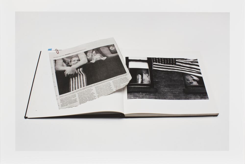 Image of a book lying open to a page featuring a photo of the American flag hanging from a building. A newsclipping featuring a photo of children behind a barricade and an adult holding an American flag lies on the book across the open page.  