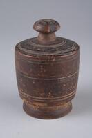 Container made by turning a single piece of wood on a lathe. Lid is crowned with mushroom-shaped knob, and shoulder is formed around mouth to support lid. Decorated with sets of double concentric circles at two places of the upper surface of the lid and at one place on the side. Around the body is carve two sets of double circles. Container is painted with red ocher.<br />
<br />
These containers were made by turning single pieces of wood on a lathe. Their lids are crowned with mushroom-shaped knobs, and shoulder is formed around the mouth to support the lid. Plate 345 (UA2004.60) is decorated with sets of double concentric circles at two places of the upper surface of its lid, and at one place around its side. Also around the body are carved two sets of double circles, and the container is painted with red ocher. Meanwhile, the lid and the body of plate 346 (UA2004.61) are ribbed. A letter resembling the Chinese character &ldquo;mok (木: wood or tree)&rdquo; is written in ink on the base of this container. 