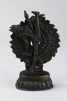 Shiva dances upon the back of a dwarf figure crouching on his stomach.  This pose is not as typical as some of his other dancing postures.  He dances on one leg extended straight down with the other straight up almost making a straight line.  His torso and head are thrown back.  He only has two hands, one holding a drum at his knee and the other a khatvanga, a club made of a bone with a skull at the top.  He wears jewelry including a belt with pendent loops and tassels, necklaces including a heavy long necklace, armlets and bracelets and an elaborate crown organized with a number of peaks. On his forehead we find the third eye, another identifier of the god Shiva. A dense circular background consisting of three inner rows of round elements, the middle one may be composed of skulls, and an outer row of flame-like elements surrounds the whole figure, animating the image into a whirl of the dance.  The whole sits on a circular base, plain with a flanged bottom section decorated with a lotus petal motif.  <br />