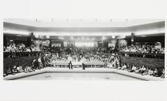 A black-and-white photograph of a crowd gathered in a food court. A panel sits at a long table, in front of which stands two men. In front of them, in the foreground, is a large pool.