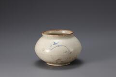 A glazed porcelain jar, whose body is wide and round with a lipped, wide opening. Detailed with a Korean character, possibly a label for the jar&#39;s contents, in blue.<br />
<br />
This voluminous jar is decorated with simplified foliage designs on the body in underglaze cobalt blue in two places. Glaze has been wiped away from the rim and the foot rim. The body features a crack and several black spots. It is made from the kaolinite-rich clay which formed thin and light walls.<br />
[Korean Collection, University of Michigan Museum of Art (2014) p.175]