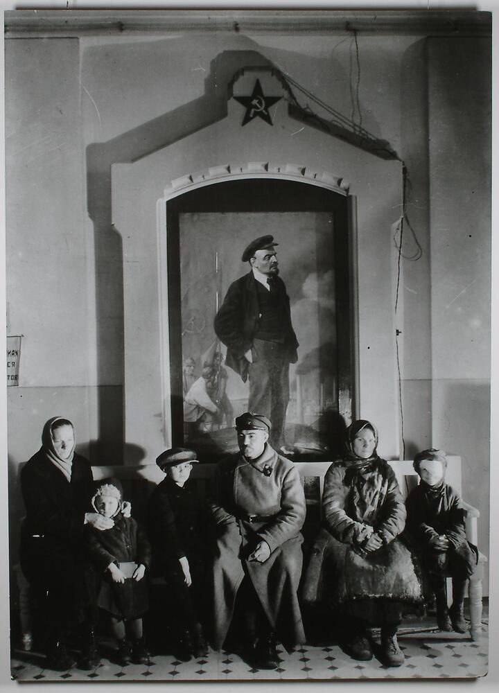 A photograph of people waiting in line to enter a children's clinic. There are two women, one man, and three children sitting on a bench in front of a large-scale painting of Lenin.