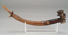 The pipe consists of a long, curved stem, a bone mouthpiece, and a bowl delicately sculpted in the form of a male head. Rectangular ears protrude sideways, while a curved chin and inverted T-shaped nose jut forward. Geometric designs characteristic of Kuba carving adorn the face, neck, and coiffure. A number of brass tacks stud the pipe, and fine copper wire has been carefully wound around the stem. Camwood powder, highly prized throughout Central Africa, has been added to its surface.<br /><br />
Reference:<br />
Maurer, Evan M. and Niangi Batulukisi.  <em>Spirits Embodied:  Art of the Congo, Selections from the Helmut F. Stern Collection</em>.  Minneapolis:  The Minneapolis Institute of Arts, 1999.