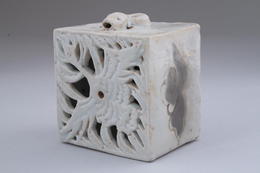 A square porcelain water dropper. The porcelain is white and there is an image of a lizard or dragon on the top. Clouds are depicted on the sides with the image of a flying crane on the front of the square. The white glaze has chipped of or is thin in places, revealing the copper underglaze.<br />
<br />
This is a cubic water dropper featuring an openwork design of a crane with wings spread on the upper face. The spout is in the shape of a newt; it is designed in the way that water drops from the mouth of the newt. The side walls feature cloud designs in copper red which were blackened during firing. The entire foot was glazed, but the glaze was wiped away from the foot, on which were placed fine sand support during firing.<br />
[Korean Collection, University of Michigan Museum of Art (2014) p.182]
