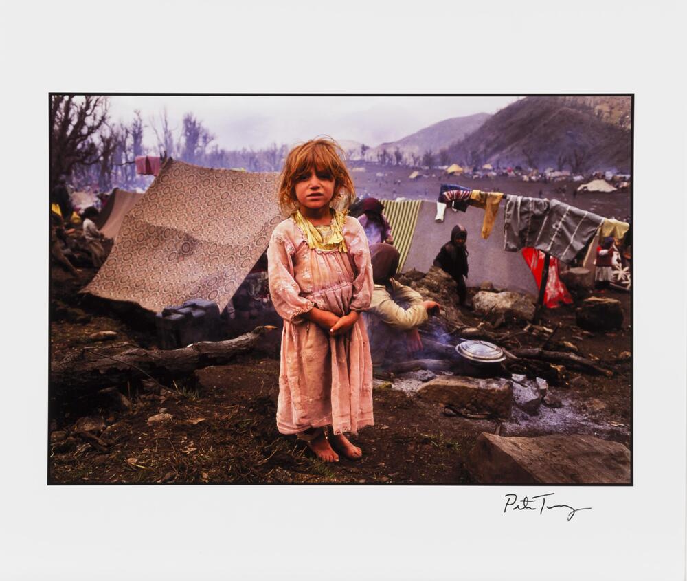 A color photograph of a young girl standing with her hands clasped, facing the viewer. She is wearing a pink dress with a yellow collar. In the background, tents and a clothesline are visible. A person cooks over a fire.