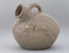 A globular pot with a flat bottom, a neck and mouth angled to one side, and a coil handle curving up from neck to top of pot.  There is bow-string decoration around pot and neck, and it is covered in a gray-green glaze. 