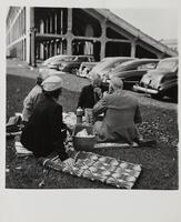 Image of two men and two women picnicking on a blanket near cars outside of a stadium.