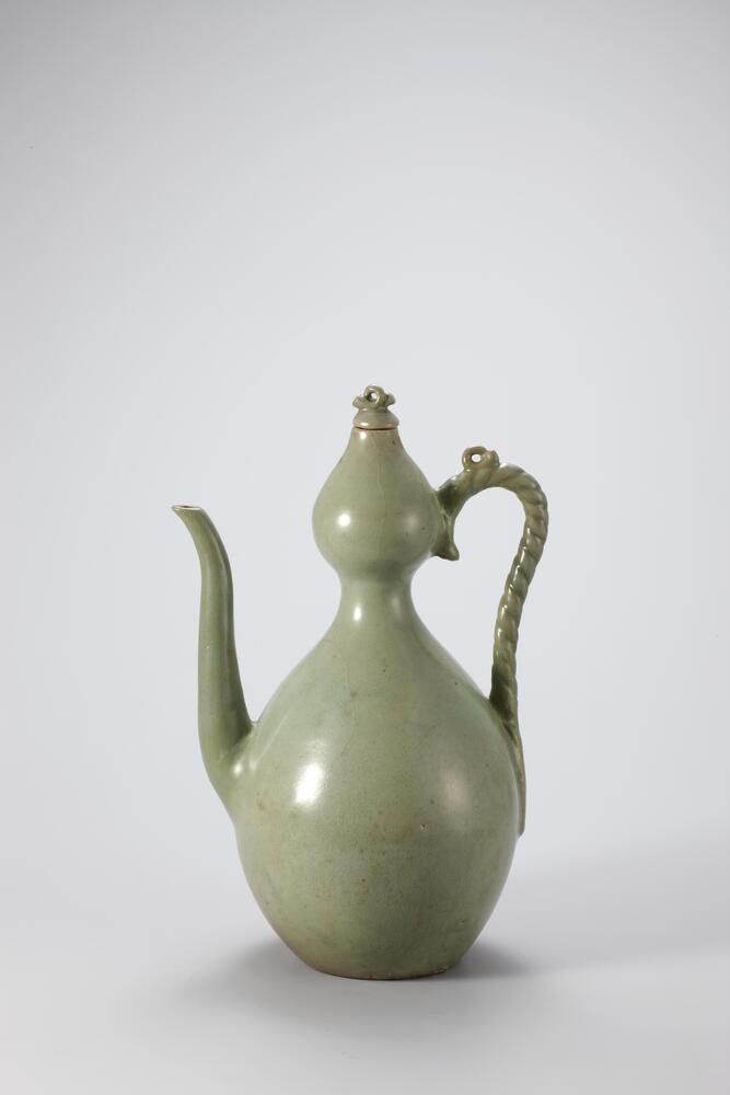 Stoneware ewer with celadon glaze in the shape of a calabash, or double-gourd. The handle mimics the tendrils of the gourd. A long, thin spout curves slightly outward from the rounded base, and a small lid crested by a tendril-like loop covers an opening at the top of the ewer.
<p>This is an undecorated gould-shaped ewer. Its stopper, handle joints, handle, and spout show traces of repair and restoration. The at base retains four refractory spur marks and was glazed only in the center of the outer base. The glaze was not fused well in parts, leaving some runnings, but it is well balanced in form.<br />
[<em>Korean Collection, University of Michigan Museum of Art </em>(2014) p.132]</p>
