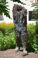 A large bronze sculpture resembling a headless and armless human figure standing with one foot forward. The surface texture is irregular and rough, with long vertical striations—resembling tree bark—and varied, colored patinas including gold, green, and whiteish gray.