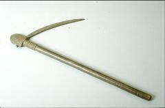 This blade has a long wooden handle. At the top of the handle is a ribbed section before an engraved top. Inserted in and protruding from the top is a long, thin blade.  