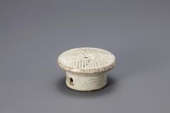 A small glazed porcelain piece resembling a cake stand. The base of the mold is wide, round and vertical rather than tapered. The top is detailed with a carved pattern that would imprint the rice cake being molded on it.<br />
<br />
Rice cake stamps are used to impress designs upon rice cakes. They are generally made of wood or ceramic. Ceramic rice cake stamps normally come in the form of round stamps and consist of a patterned surface and a handle. Patterns, carved or raised, on the stamp vary from geometric lines to auspicious designs that wish for prosperity and longevity. Their small size makes them highly portable, while their simple yet contemporary designs have mad them popular among collectors. The University of Michigan Museum of Art collection includes nine white porcelain rice cake stamps. Some are gifts from Mr. and Mrs. Hasenkamp, and others are gifts from Ok Ja Chang and the Chang family.<br />
<br />
This rice cake stamp features a geometric line design. Its walls are thick and heavy. The