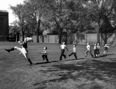 A black and white image of a man in a uniform marching across a lawn followed by six young children in a line.