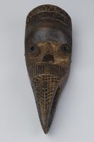 Bird mask with an elongated beak that is decorated with a carved geometric design. The mask has a protruding nose, forehead, and eyes. The top of the mask has a carved geometric design and a bent nail embedded into the top.&nbsp;