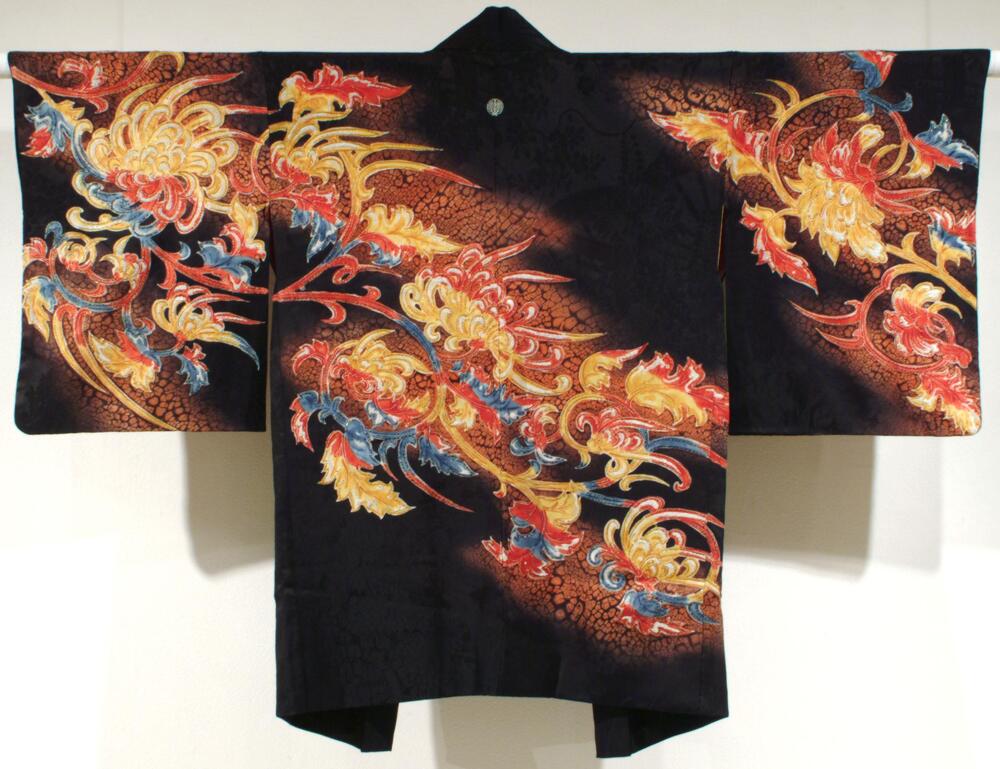 It is a black silk damask haori (short jacket for kimono) with wax-resist patterns, hand-painted design and metallic threads embroidery. The haori is in medium length, covering just underneath hip. It has elongated sleeves. The silk fabric is woven in a twill pattern of palace carts and flower baskets. Then the fabric is dyed with black. The white family crest under the collar and the diagonal part where chrysanthemum design would appear are left out from dying. The pinkish orange scale pattern is added using wax-resist technique. Chrysanthemum design is hand-painted with white, red, yellow, and blue colors. Finally embroidery is added in various metallic threads around the contours of the chrysanthemum petals and leaves.<br />Orange satin damask lining with woven wave design, with stenciled (?) designs of white flying cranes.  Silver cord on one side, gold on the other, both with tassels.
