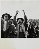 Image of a large crowd of people. In the center foreground is a man wearing a long jacket and round-rimmed hat with his arms raised in the air and his mouth wide open.&nbsp;