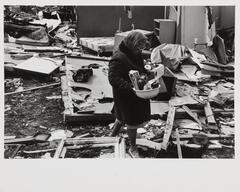 A black and white photograph of a woman holding a caulinder full of objects. She walks through an area filled with scattered, broken furniture, windows and walls.
