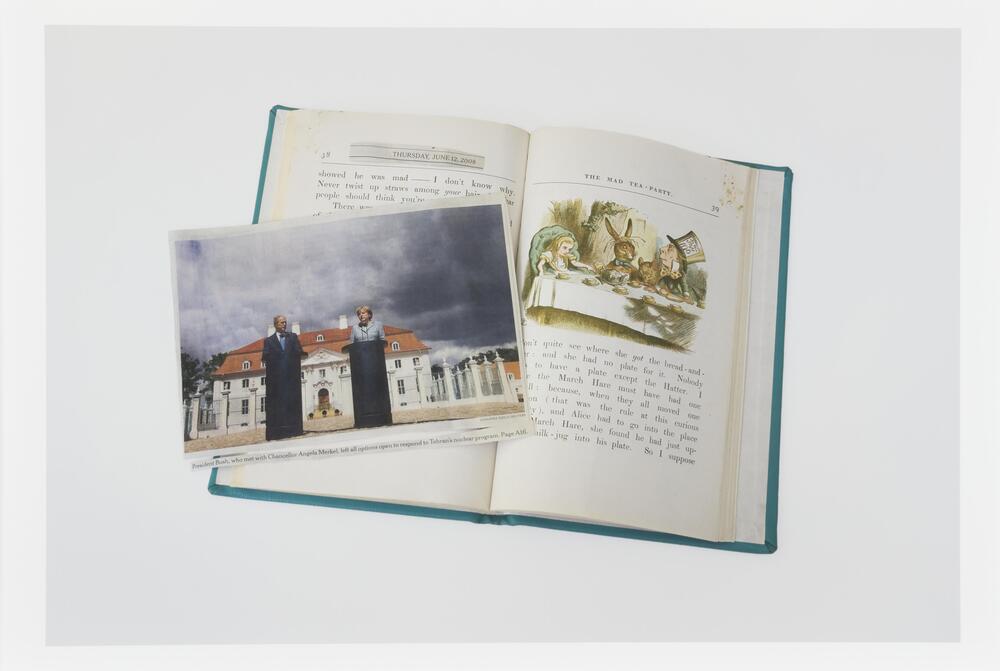 Image of a book lying open to a page featuring an illustration of the Mad Hatter's tea party from Alice in Wonderland. A newsclipping featuring a photo of George W. Bush and Angela Merkel lies on the book across the open page.  