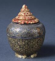 The small container with lid has a deeply engraved design filled with black enamel. The body has tight, compact, overall decoration of floral motif, with the stupa-shaped gold fitting on the top. Colored glass pieces were inlayed in the fitting.<br />