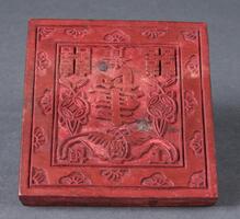Wooden block painted cinnabar red with a carved floral design framing an inner square that contains six characters in the top and lower right, center, and left corners and a large character in the middle (“longevity”) with an image of two coins suspended from the wings of a stylized bat in the lower center.<br />