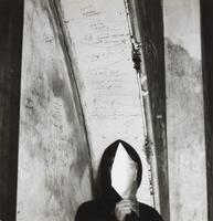 A photograph of the bust of a young man, cloaked in black, holding a reflective shard of glass in front of his face. He stands in front of a wall of graffiti.