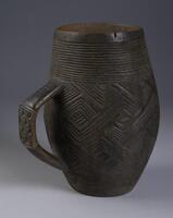 Vertically positioned cup with a slight bulge in the middle. Trapezoid shaped handle with intersecting linear design on it. The top and base of the cup are carved with multiple horizontal lines and in between are interlocking chevrons.