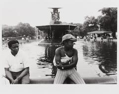 Photograph of a man sitting on the lap of a woman on the ledge of a public fountain.